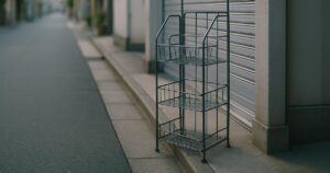 An empty wire newspaper rack beside a closed shop shutter on a quiet Tokyo side street at dawn